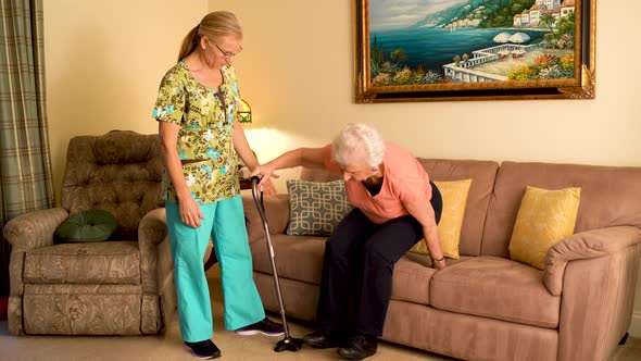 Home healthcare nurse helps elderly woman get up from couch and use a cane. alt