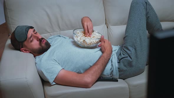 Tired Man Relaxing on Sofa in Front of Television Eating Popcorn While Watching Movie Show alt