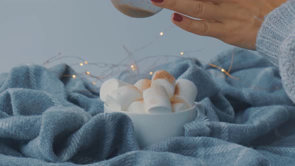 Adding Cacao Powder on Top of Hot Chocolate with Marshmallows in White Cup on Sweater Background alt