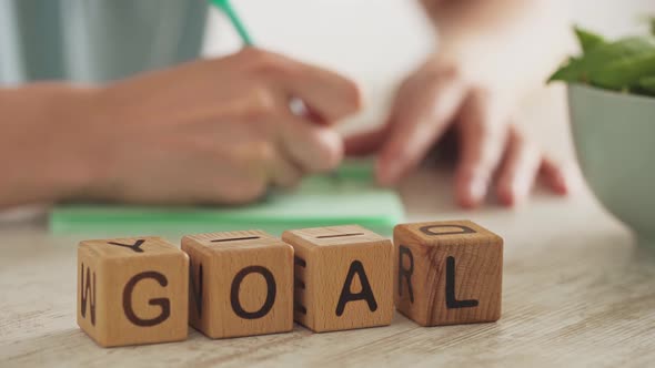 The Word Goal Is Assembled From Wooden Cubes. In The Background, A Female Hand Writes Down The Goal alt