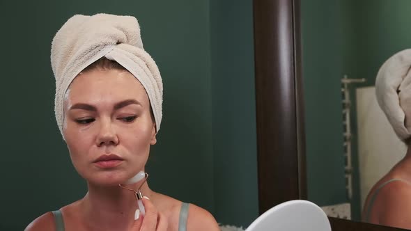Young Woman Using Jade Facial Roller for Neck Massage Sitting By the Bathroom Mirror Slow Motion alt