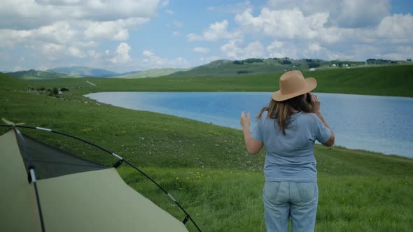 Back view of millennial woman in hat and  blue t-shirt drinking hot tea from thermos alt