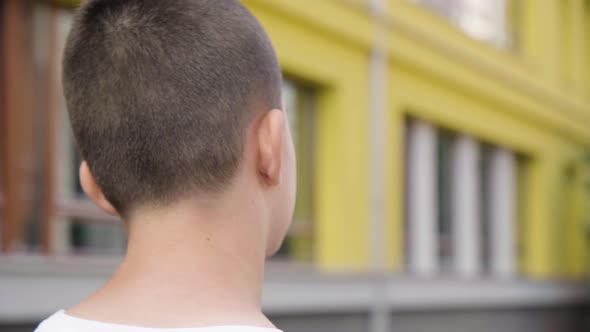 A Caucasian Teenage Boy Looks Over His Shoulder at the Camera  Closeup  a School in the Background