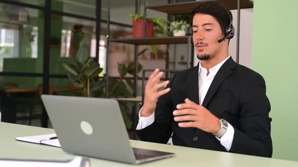Friendly Latin Male Office Employee with a Headset Using Laptop for Communication with Coworkers