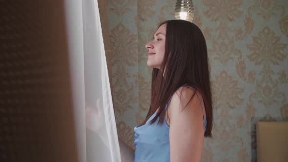 Young Woman is Standing Near the Window Stroking Long Dark Hair with Hands and Enjoying Sunrise