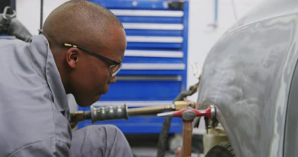 African American male car mechanic holding a screwdriver and using a hammer to repair a car alt