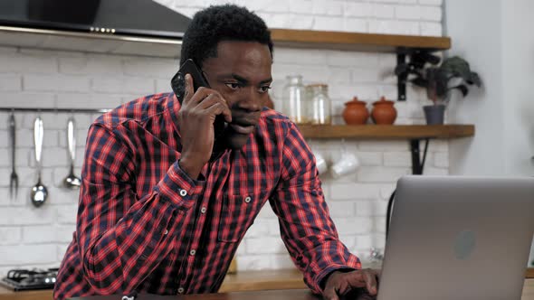 African American Man Uses Laptop Talking on Mobile Phone Standing in Kitchen alt