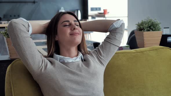 Serene Young Woman Resting on Couch Holding Hands Behind Head Taking Deep Breath of Fresh Air. alt
