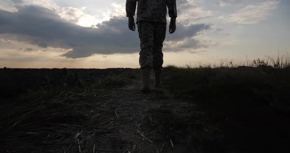 A Soldier in Military Uniform Walks Across a Black Burnt Field at Sunset alt