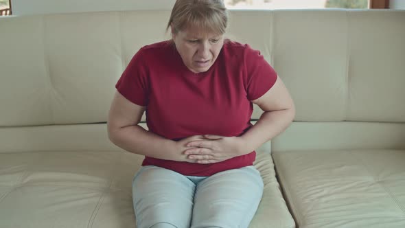 Portrait of a caucasian woman with blonde hair sitting on sofa having strong stomach ache alt