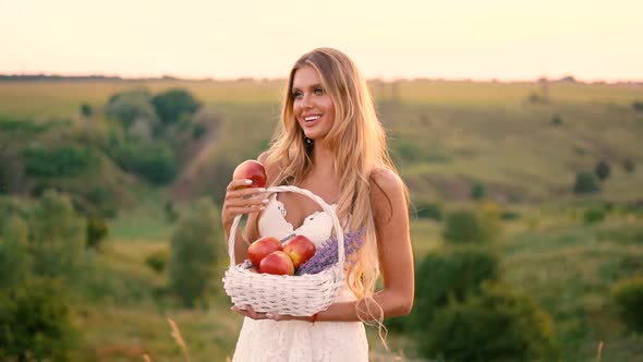 Beautiful sexy blonde girl in white dress posing in a field at sunset with a basket of fruit
