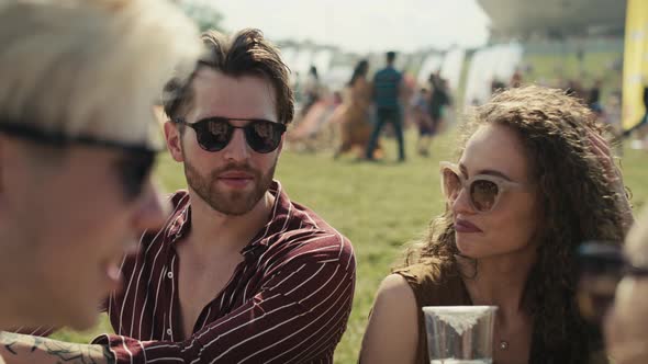 Group of friends sitting on grass together at music festival and drinking beer while chatting. Shot alt