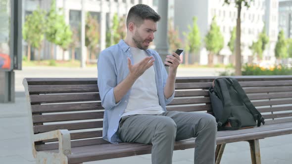 Young Man Reacting to Loss on Smartphone While Sitting on Bench alt
