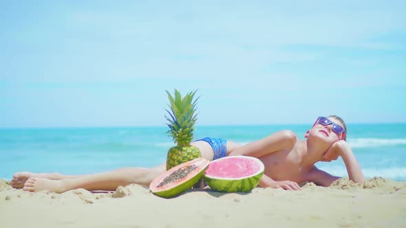 Boy Lies on the Beach with a Composition of Exotic Fruits Against the Sea alt