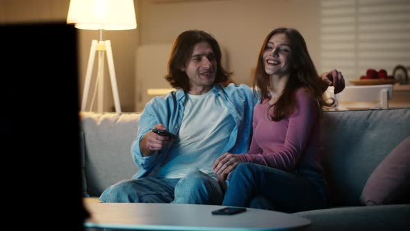 Happy Young Couple Sitting Down on Couch at Home Embracing and Choosing Program on Tv Slow Motion alt