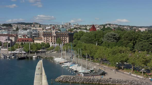 Drone flying backwards from small yacht harbor near city revealing  sailboat leaving the harbor alt