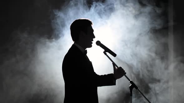 Showman Entertains Audience at Microphone in Light of a Searchlight Among Smoke alt