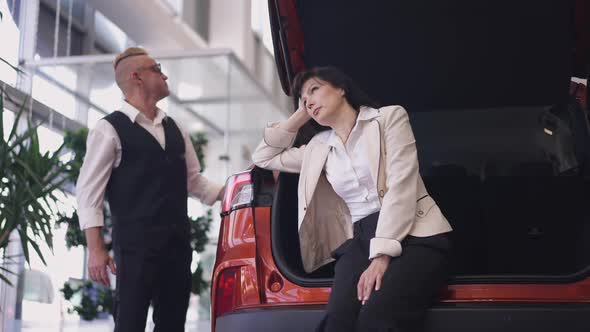 Portrait of Bored Woman Sitting in Car Trunk Sighing As Man Choosing New Vehicle in Dealership in alt