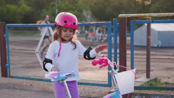 Cute Light Hair Little Girl in Pink Helmet in Elbow and Knee Pads Sits on Bicycle