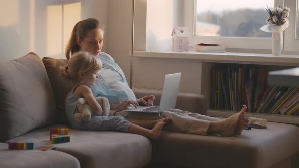 Pregnant Woman and Little Girl Relaxing on Couch with Laptop alt