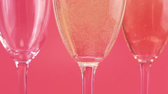 Sparkling wine is poured into glasses on a pink background with heart-shaped confetti. alt