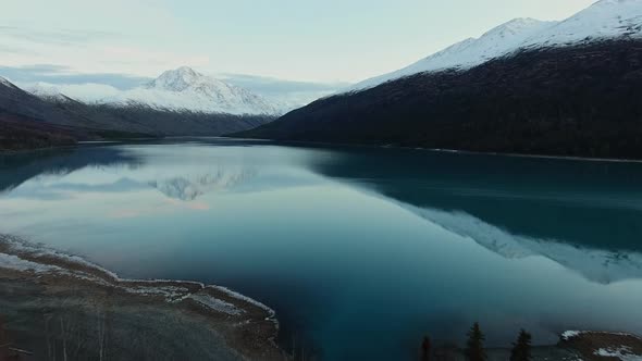 Drone approaching the snow-capped mountains reflected in a calm river in Eklutna Lake, Alaska, USA alt