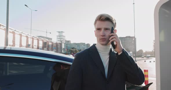 Businessman Talking On The Phone Leaning On A Blue Luxury Electric Car Which Is alt