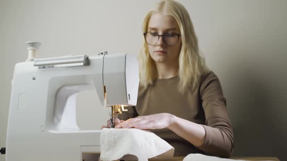 A Young Woman Sews Clothes Sitting in a Light Workshop at a Table with a Sewing Machine alt