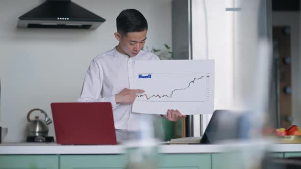 Confident Asian Man Pointing at Graph Talking Looking at Web Camera on Laptop alt