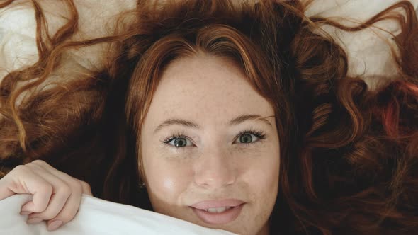 Attractive Caucasian woman with red hair playfully looks out from under blanket