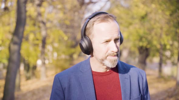 A Middleaged Caucasian Man Listens to Music with a Headset and Walks in a Park in Fall with a Smile