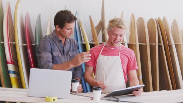 Two Caucasian male surfboard makers working on projects using a laptop computer alt