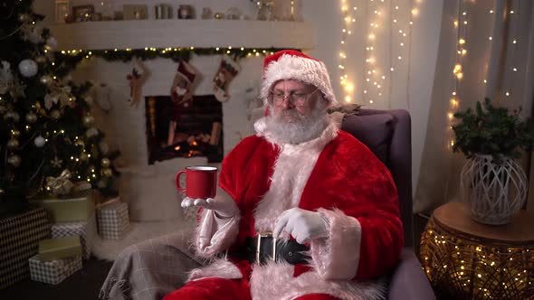 Funny Santa Claus Sits in a Chair and Jokes with a Cup Near the Christmas Tree and Fireplace in the