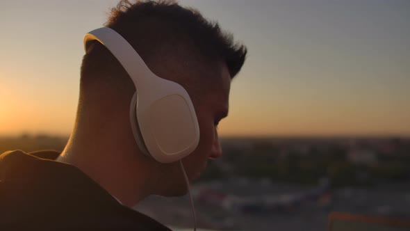 Close-up of a Man in Headphones Listening To Music on the Roof at Sunset with a View of the City. alt