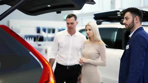 Car Agent Showing Trunk to Customers Car in Automotive Dealership Showroom alt