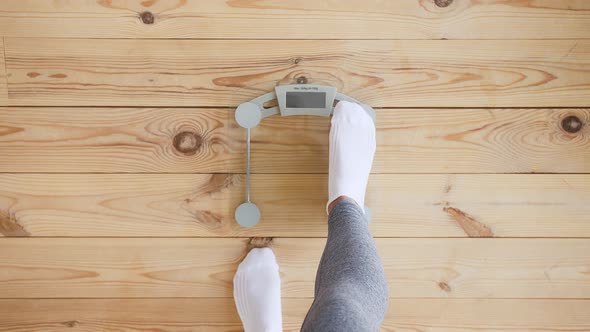 Unknown Female in Gray Leggings and White Socks is Standing on Weight Scale on Wooden Floor alt