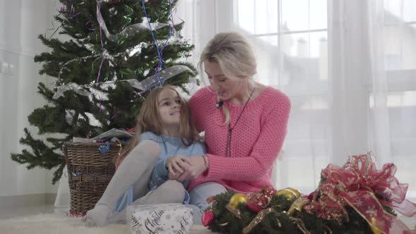 Smiling Caucasian Girl Sitting with Adult Woman at the Background of Christmas Tree alt