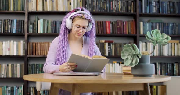A Young Woman in Headphones with Long Colored Hair Sits at a Table with a Laptop and Houseplant
