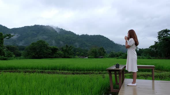 A young woman drinking coffee while looking at a beautiful rice field and nature view