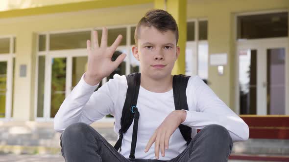 A Caucasian Teenage Boy Waves at the Camera with a Smile As He Sits in Front of School  Closeup