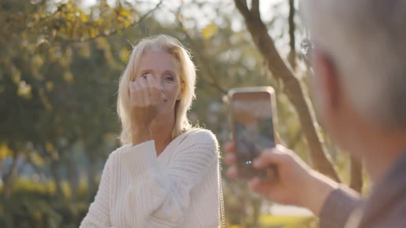 Male Pensioner Taking Picture of Attractive Senior Woman on Smartphone in Park alt