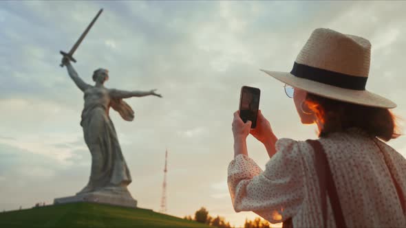 A young tourist in a hat taking a photo of the Motherland on a smartphone on the Mamayev Kurgan alt