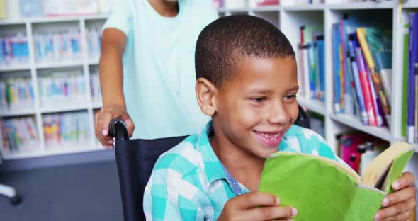 Happy schoolboy carrying his friend in wheelchair at library alt