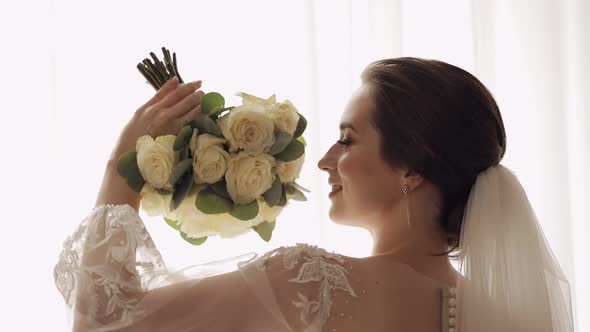 Bride in White Dress Staying Near Window with Flowers Bouquet Wedding Morning Preparations at Home alt