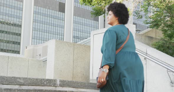 Happy plus size biracial woman walking on stairs with smartphone in city alt