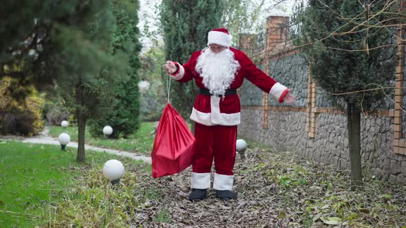 Wide Shot Santa with Gifts Bag Standing on Backyard with Green Trees alt