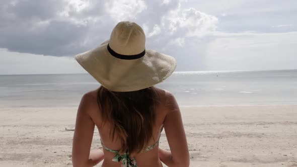 Camera following young brown haired woman with bikini and hat walking on beautiful beach towards cle alt