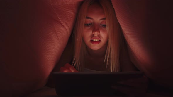 Talented Woman is Lying on Bed Under Blanket Drawing with Stylus on Digital Tablet and Smiling alt