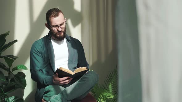Bearded Young Man Sitting Near Window and Reading a Book at the Sunrise alt