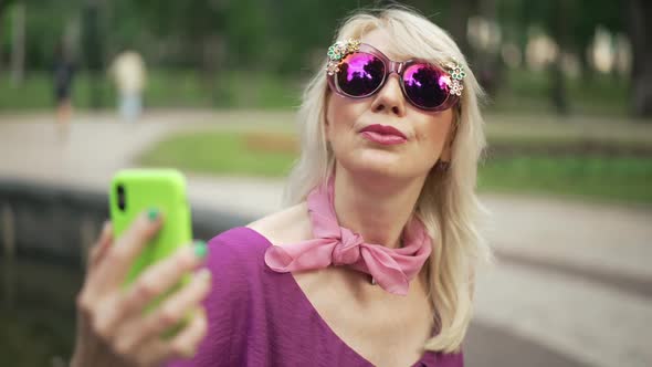 Portrait of Confident Adult Woman in Sunglasses Taking Selfie on Smartphone Resting in Summer Park alt